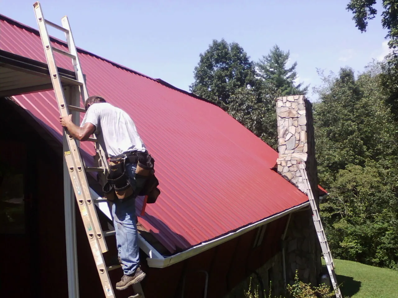 Roofer on a ladder beside a red metal roof and stone chimney — Soffit & Fascia Repair by Roofing Brothers in Mekinock